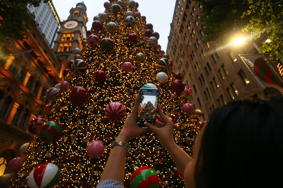 A woman takes a photograph of a huge Christmas tree outside tall buildings.