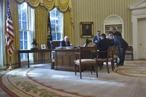 President Donald Trump speaks on the phone from the Oval Office as staffers look on.
