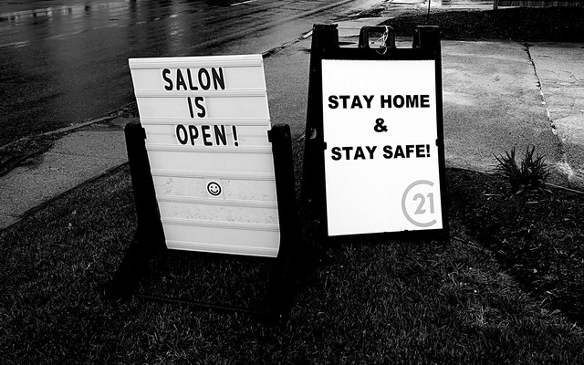A photo of two road-side signs. One says, "Salon is open!" The other says, "Stay home and stay safe!"