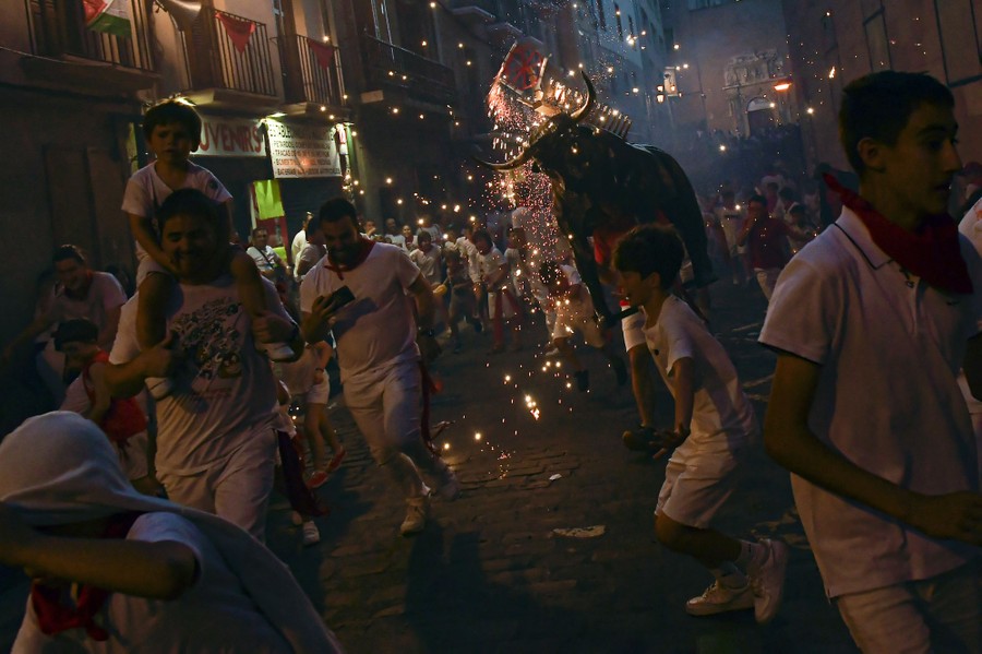Adults and children run in a street at night, playfully being chased by a person carrying a statue of a bull that is shooting sparks.