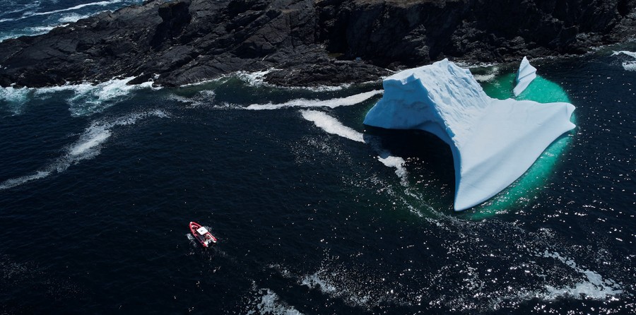 An aerial view of a small boat passing by a large iceberg near a rocky shore