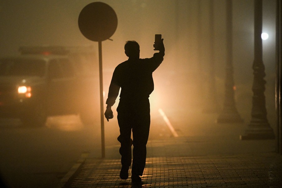 A person walks on a sidewalk during a dust storm, seen in silhouette in front of car headlights.