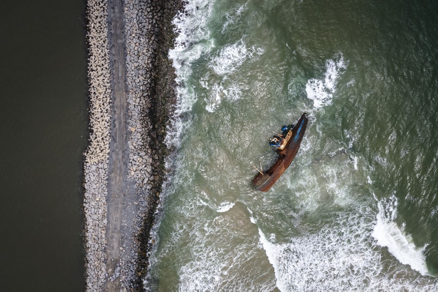 An aerial photograph of a stranded boat in crashing surf, near a breakwater