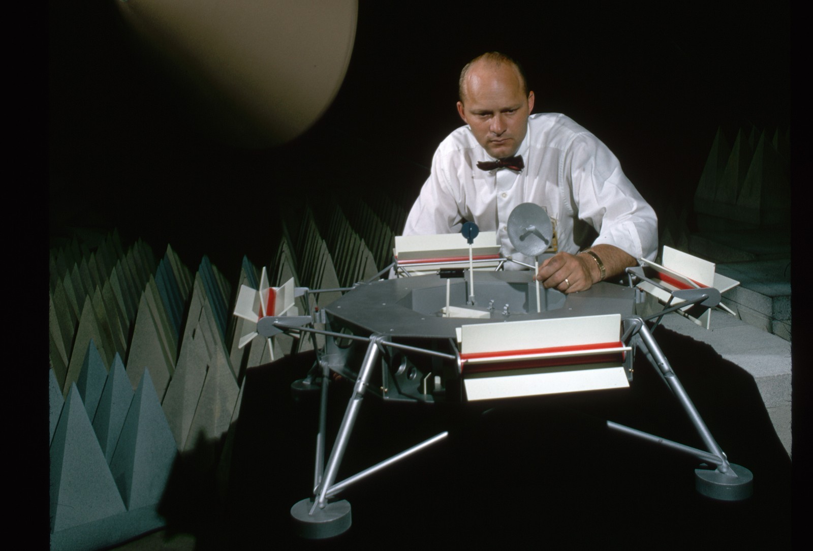 A man sets up a model of the Mars Viking spacecraft in what appears to be a sound-dampening room with conical baffles behind him.