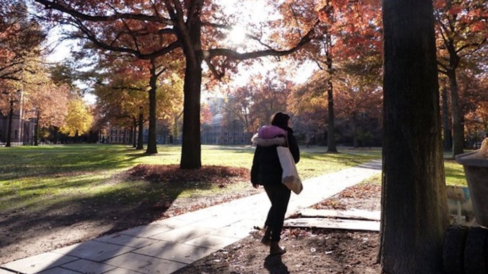 A woman walks along a sidewalk among trees in autumn.