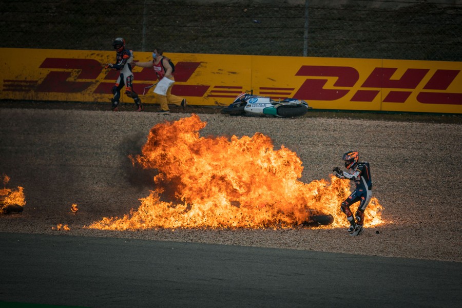 A motorcycle rider scrambles away from a fireball engulfing a motorcycle on the side of a track.
