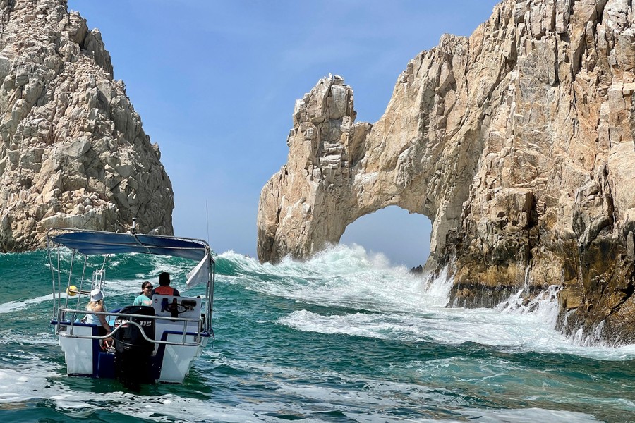 Tourists ride in a boat near waves crashing against a natural arch formation in rocks.