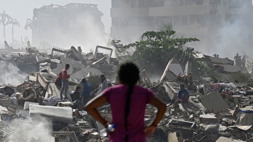 Color photograph of a woman, seen from behind, standing with her hands on her hips and looking at people digging through the rubble of a collapsed building.