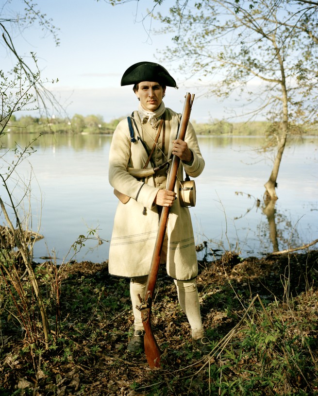 photo of reenactor as American soldier standing in black hat and tan coat with musket on the shore of Lake Champlain