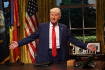 A man dressed as Donald Trump, in a long red tie with messy blond hair, holds out his arms behind a desk.