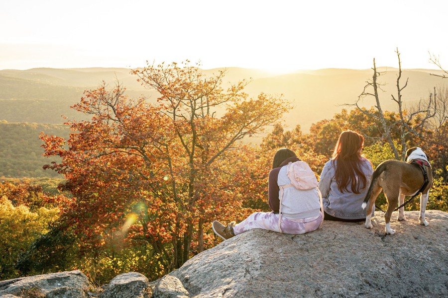 Two people sit on a large rock overlooking rolling hills, watching a sunset.
