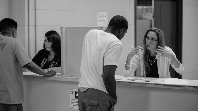 An employee of the the Sheriff's Office of Mental Health Policy and Advocacy, right, speaks with a detainee at the Cook County Jail. The black and white photo shows the employee motioning and the man leaning in to hear her.