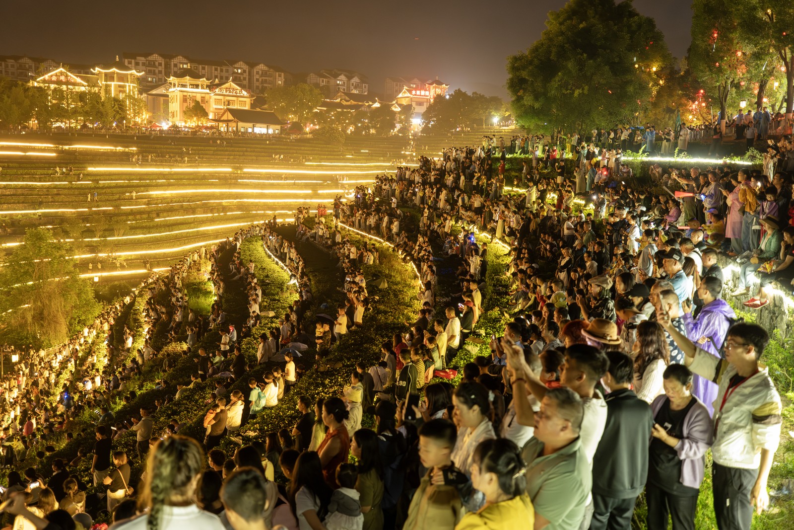 People stand in long rows along curved terraces on a hillside, watching a concert below at night.