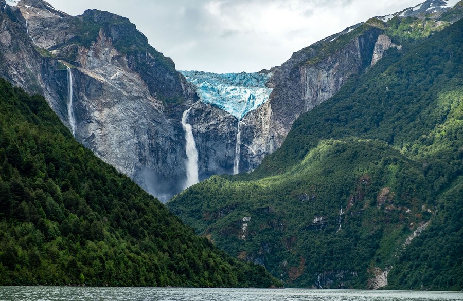 A dramatic view of steep mountains rising from water, with waterfalls along sheer cliffs beneath the visible face of a glacier