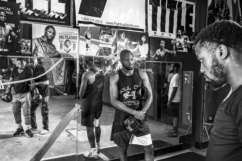Conwell in a training gym, wearing boxing gloves, stares at himself in a mirror while his trainers look on