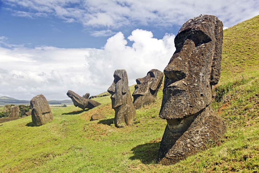Tall stylized stone statues of human heads stand on a steep and treeless mountain slope.