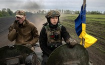 Two members of the Ukrainian 28th Mechanized Brigade ride in an armored vehicle towards hostile Russian forces. The one on the left is smoking a cigarette, the one on the right is scowling, and to their side is a blue and yellow flag.