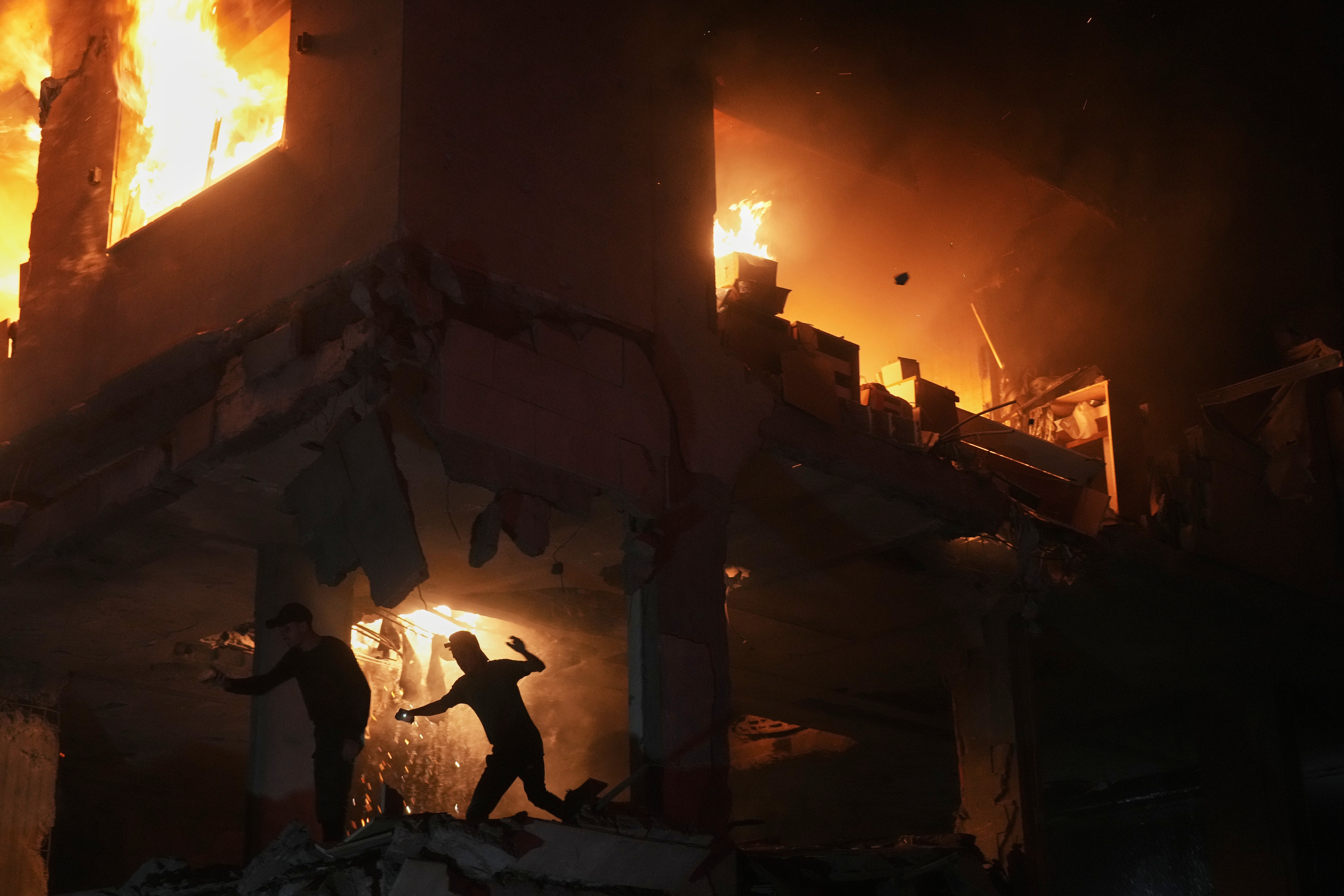Several people move beside a heavily-damaged building that is burning after a military strike.