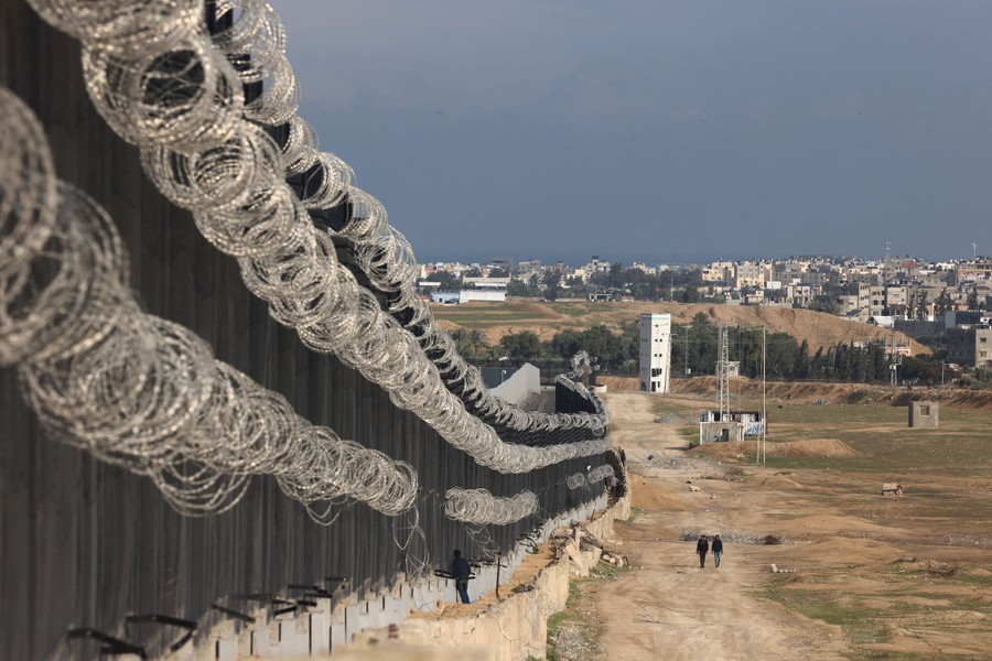 People walk beside a large border fence topped with razor wire.