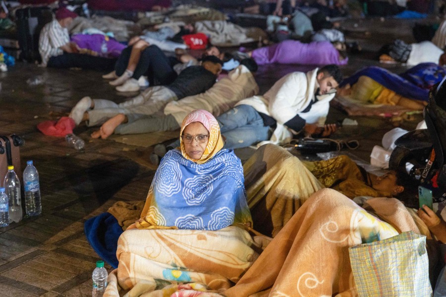 More than a dozen people sit and lie on the ground in a public square at night, wearing coats and blankets.
