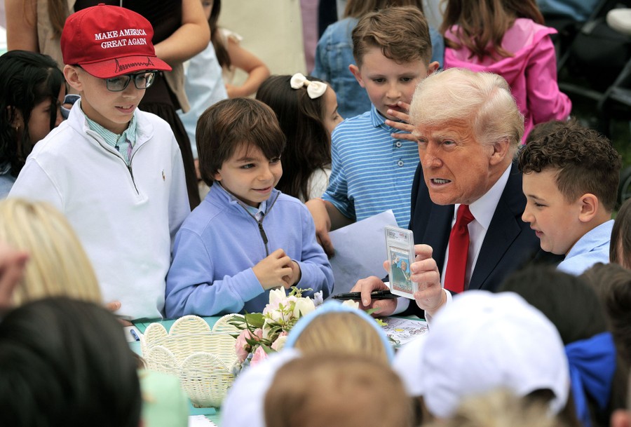 Donald Trump holds up a collectors card depicting his assassination attempt, at a table surrounded by children.
