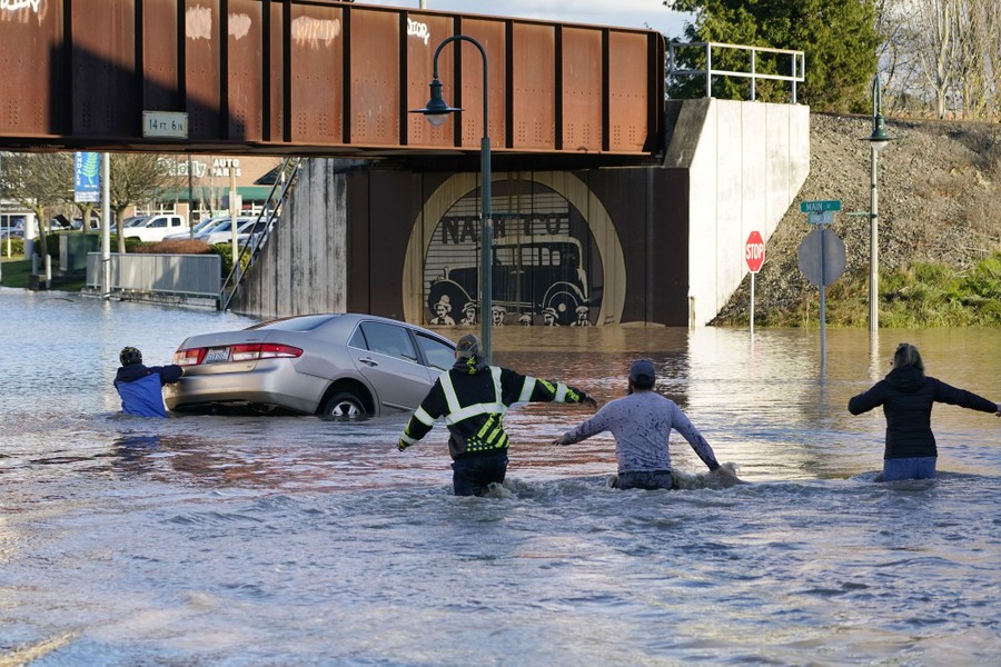 Several people wade into waist-deep water covering a road to push a partially-submerged, partially-floating car to safety.