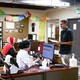 A man with a stethoscope around his neck stands in front of people sitting at computers in a doctor's office.