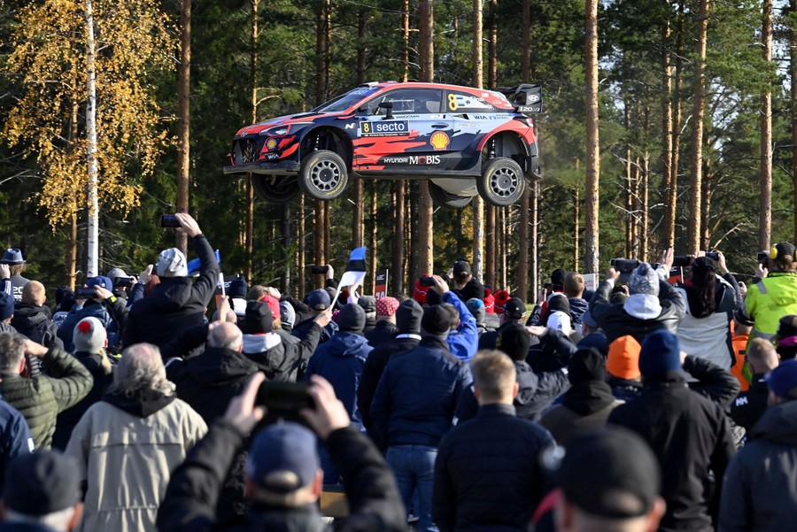 Spectators watch as a rally car flies past with a forest in the background.