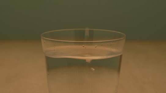 A short glass of water, resting on a light brown table, with an olive green wall in the background.