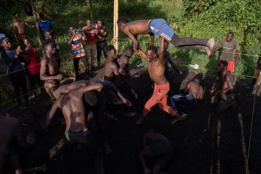 About a dozen young men scuffle in an outdoor dirt-floor wrestling ring. One holds another above his head, tossing him.