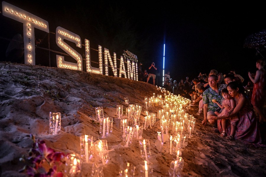 Many people gather and place candles on a beach, near an illuminated sign that reads "Tsunami."