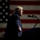 Donald Trump speaks into a microphone. An American flag hangs behind him.