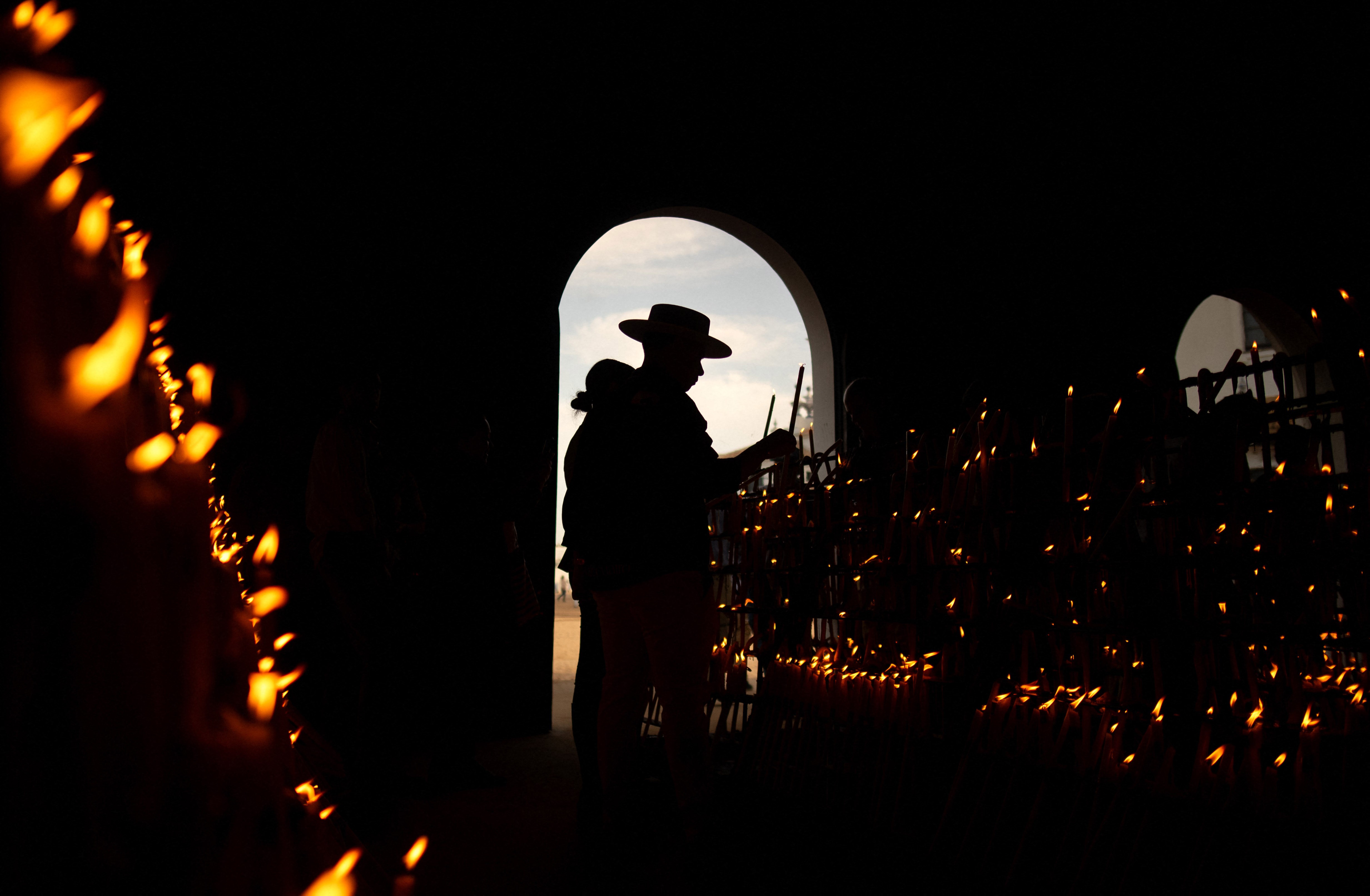 A pilgrim lights a candle in a dark space filled with many lit candles.