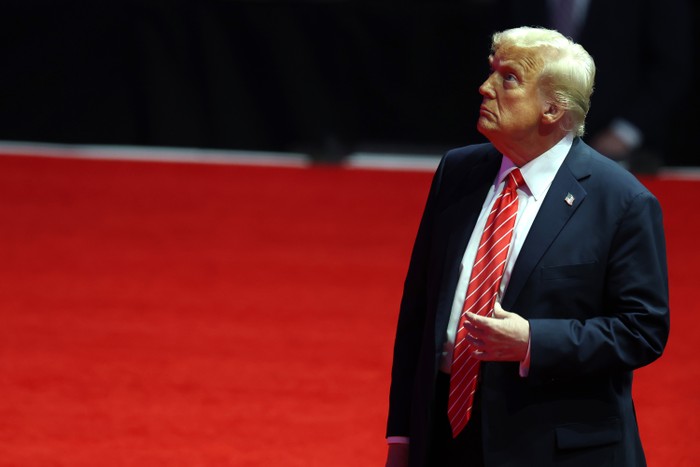 Donald Trump touches his tie and looks up while standing on a red carpet