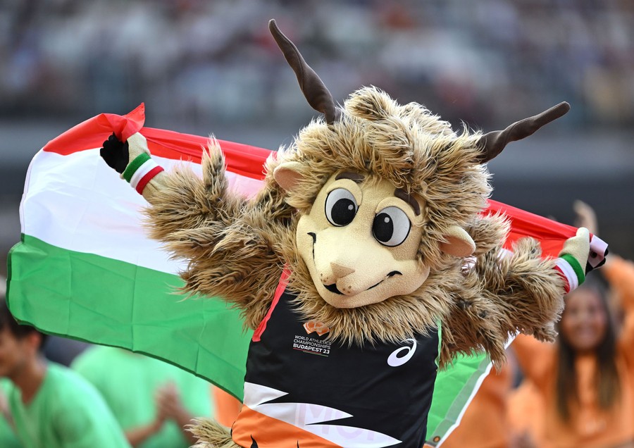 A person wearing a mascot costume, looking like a cartoon sheep, runs while holding a Hungarian flag.