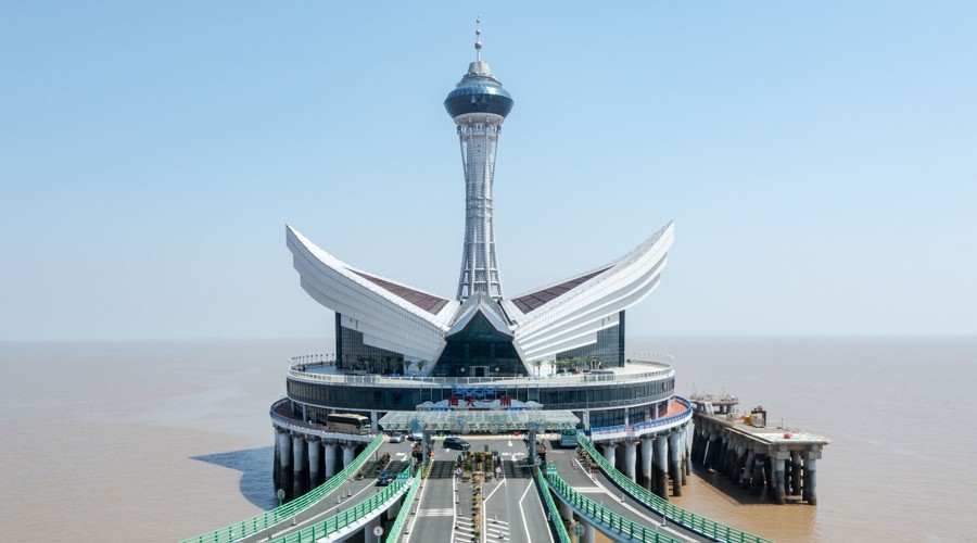 A circular service center with a stylized roof and tower sits on pilings, alongside a highway bridge over a broad bay.