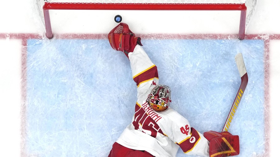 An overhead shot of a hockey goaltender reaching for—but just missing—a puck headed into the net.