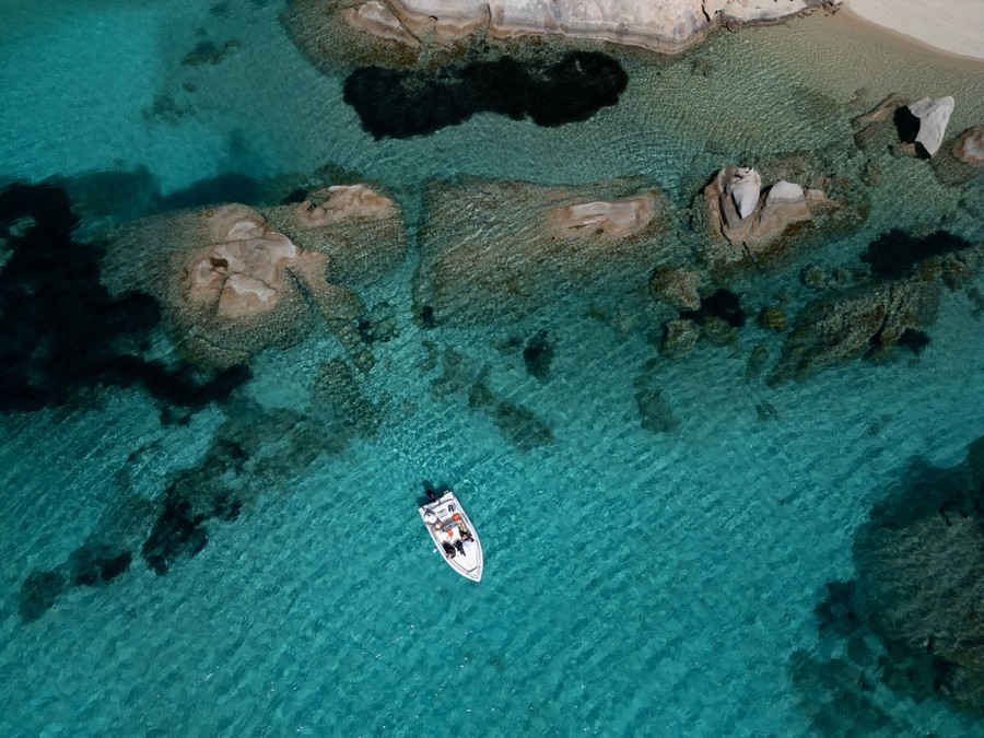 An aerial view of a small boat floating in clear blue water near a rocky shore.
