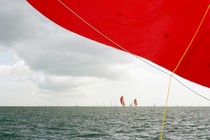 Boats with bright red sails on the ocean, one sail close up to the camera and covering the top half of the frame