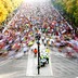 A wide street full of runners, splitting around a fork in the road. The runners in front are moving so quickly that many of them are blurs.