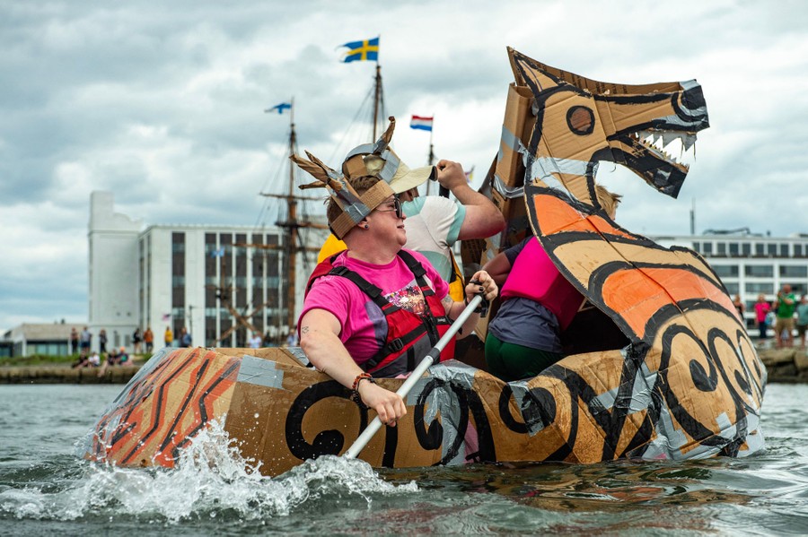Several people paddle a makeshift viking boat built out of cardboard boxes and duct tape.