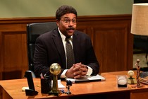 Kenan Thompson, wearing oversize glasses and a mustache, sits behind a desk with hands folded.