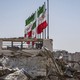 A color photo of Iranian flags flying above a bomb-damaged building.