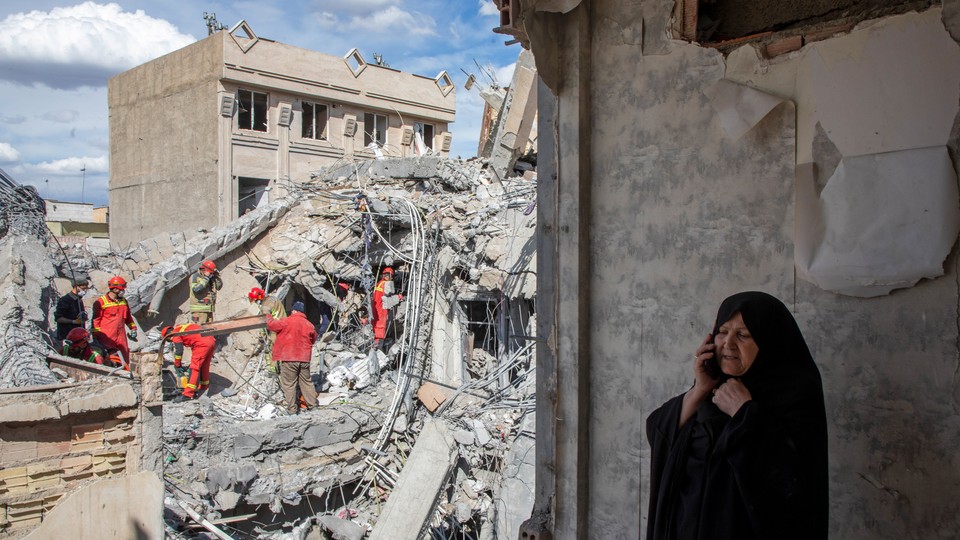 An image of a woman speaking on the phone as emergency workers sift through rubble of a residential building that was hit in an airstrike in Tehran.