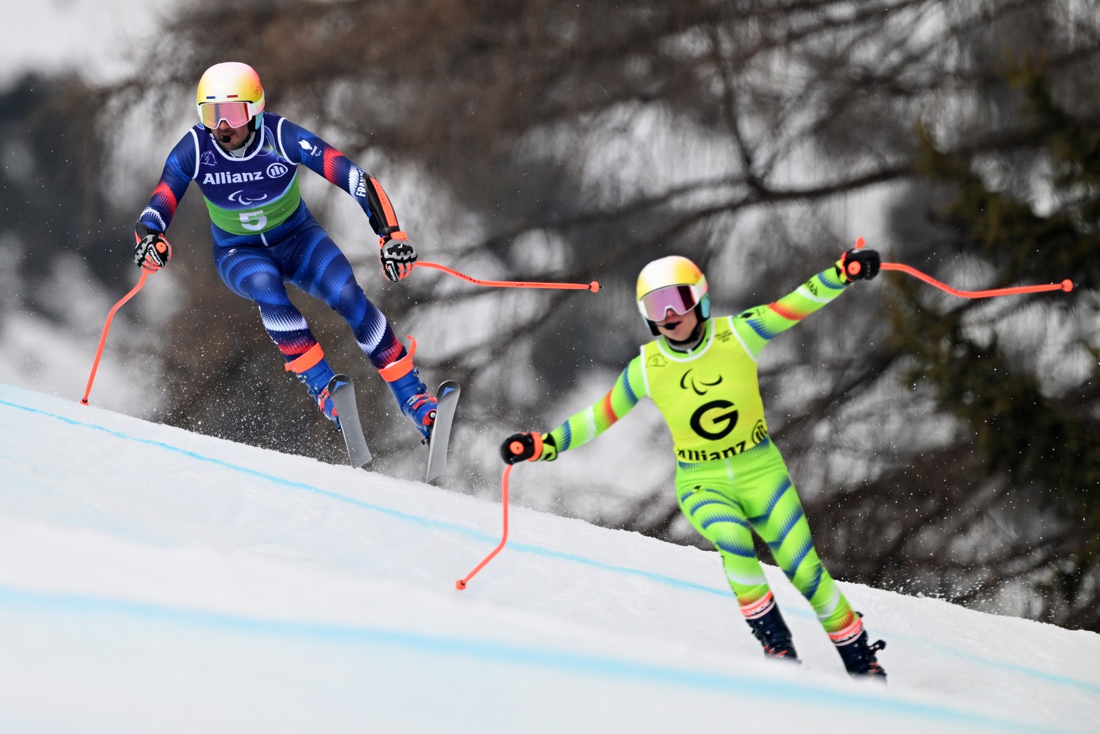 Two downhill skiers race down a course, the one in front, acting as a guide, wears a bib with a large letter 'G.'