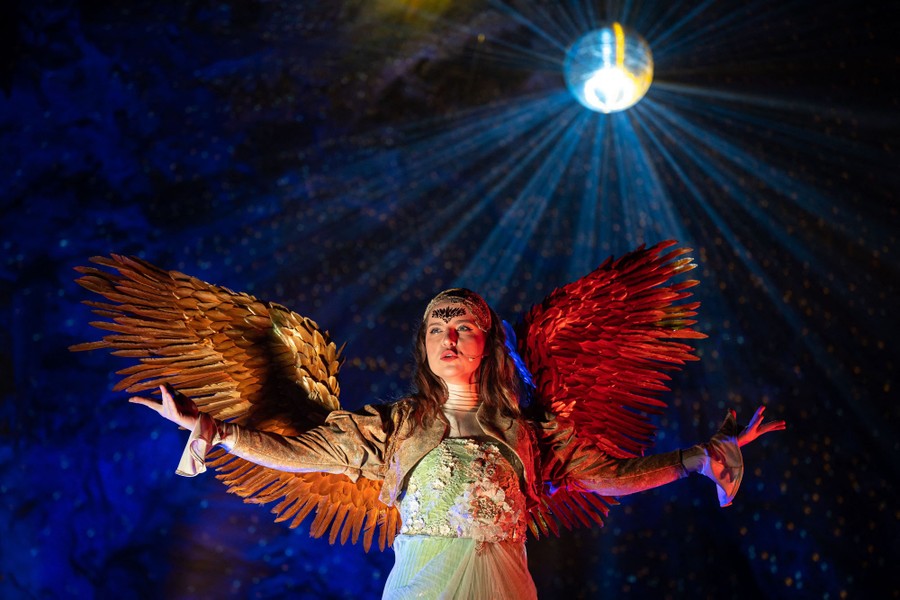 A singer in a winged costume performs beneath a mirror ball in cave.