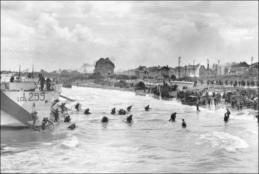 Soldiers wade from landing craft to a nearby beach, some carrying bicycles as well as weapons and packs.