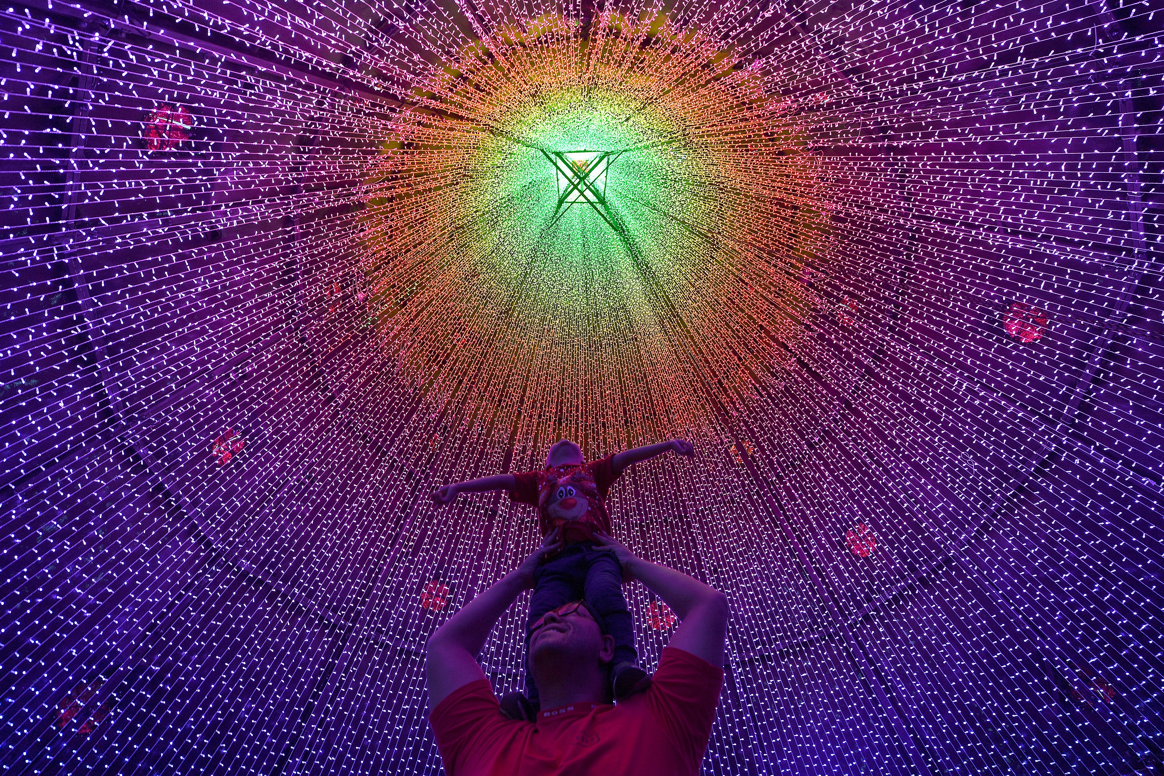A man holds a small child high on his shoulders, posing beneath a tent of colored strings of Christmas lights.