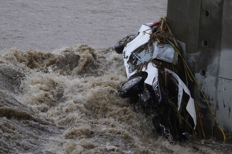 A smashed car is pressed against a concrete pillar by fast-moving water.