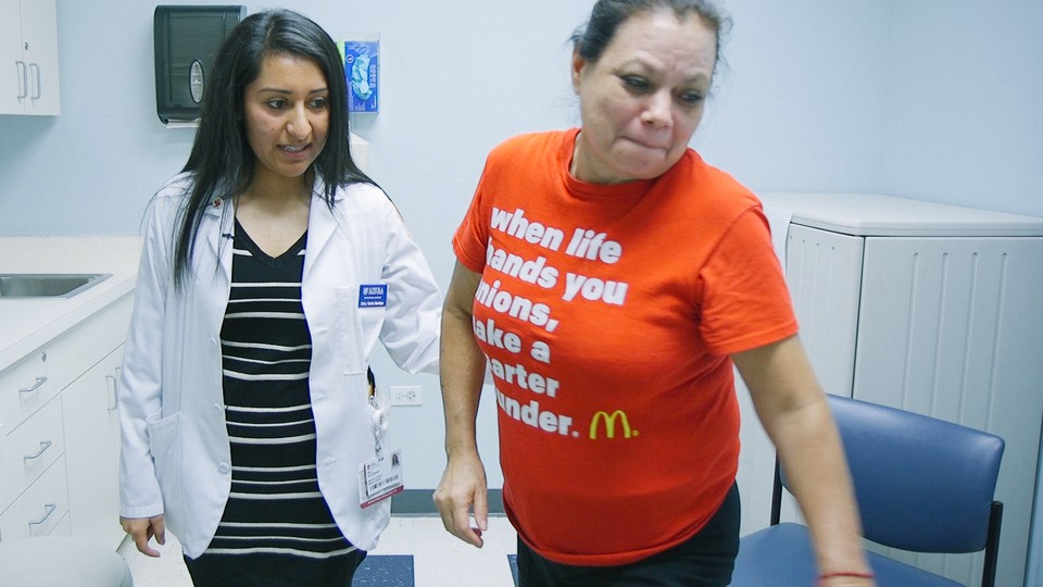 A woman in a lab coat supports a woman in a T shirt in a doctor's office.